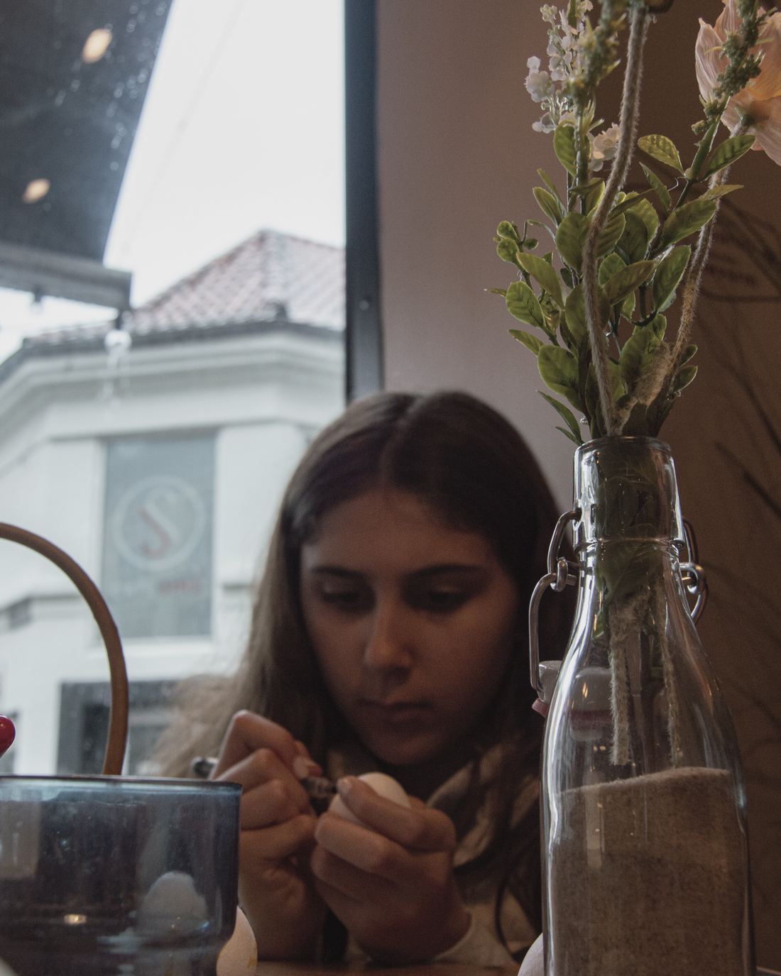 A girl painting an egg at a cafe window on a rainy day
