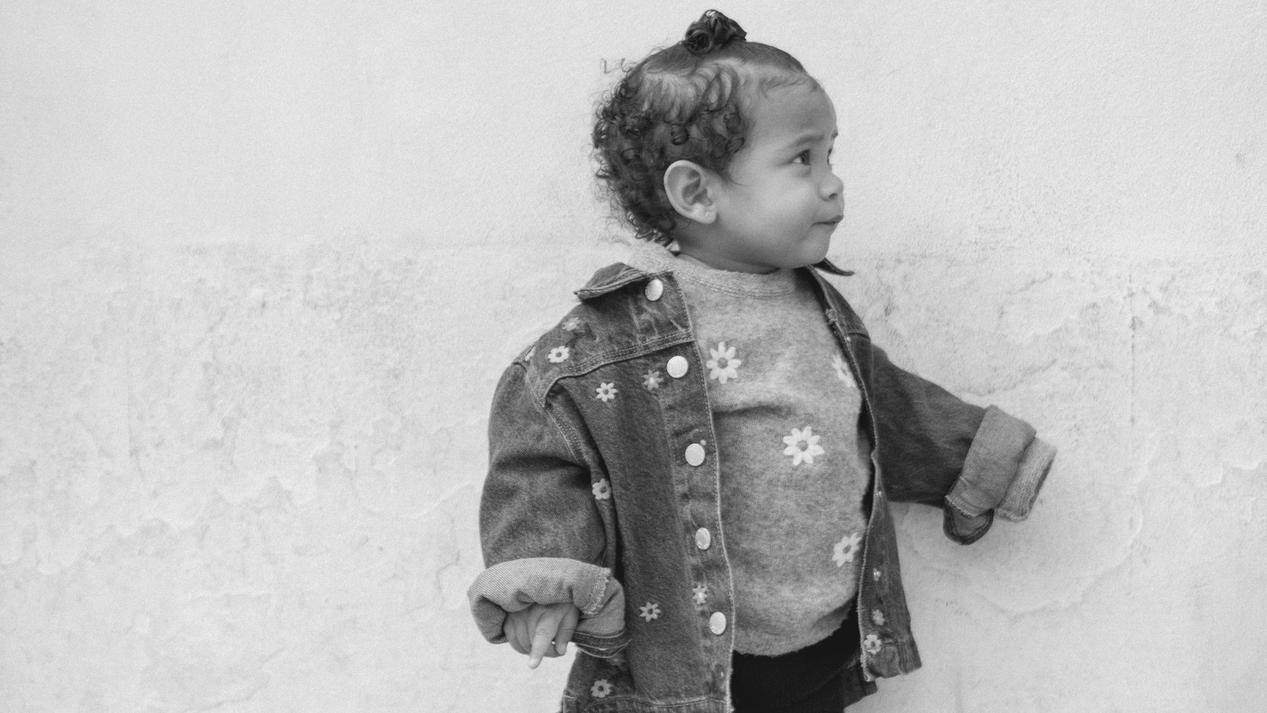 A small child in a denim jacket standing against a white wall